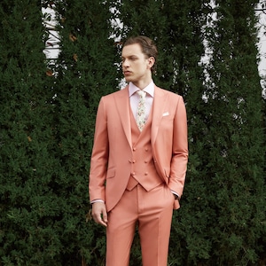 Puede incluir: Un traje de color rosa coral con un chaleco de doble botonadura y una camisa blanca con una corbata de estampado floral. El traje lo lleva una persona de pie frente a un seto verde.