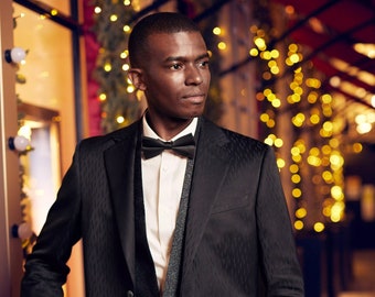May include: A man wearing a black tuxedo with a black bow tie and a white shirt. He is standing in front of a blurred background of lights.