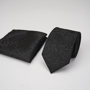 Op de afbeelding: Een zwarte stropdas en bijpassende pochet set. Beide items hebben een terugkerend, wervelend patroon. De stropdas is geknoopt en gedeeltelijk opgerold, de pochet is netjes gevouwen. De set ligt op een witte ondergrond.