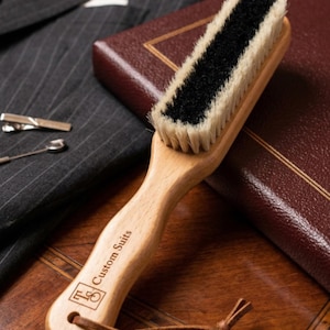 May include: A wooden clothes brush with a black bristle center and light bristles. The handle is engraved with "Custom Suits". A dark gray pinstripe suit, a leather-bound book, and accessories are in the background.