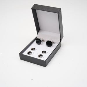 May include: A set of two round silver-toned cufflinks and four matching round silver-toned shirt studs, each featuring a smooth black circular inlay. The items are presented inside a black rectangular hinged display box with a white interior.
