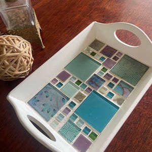 May include: White rectangular tray with a mosaic tile design. The mosaic features squares of teal, purple, and green glass tiles with patterned accents. The tray has two handle cutouts and is on a wooden surface.