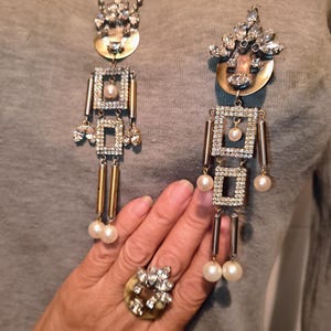 Può includere: Un set di gioielli ornati composto da collana, orecchini e anello abbinato. La collana e gli orecchini presentano un design geometrico con cristalli trasparenti, perle finte e dettagli in metallo dorato. L'anello ha un gruppo di cristalli.