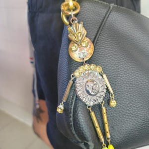 Può includere: Un ciondolo decorativo per borsa con un design a forma di volto di sole. Il ciondolo ha una clip e dettagli in metallo color oro, con un ciondolo a forma di volto di sole color argento. Perline gialle e verdi pendono dal basso. Il ciondolo è attaccato a una borsa nera.