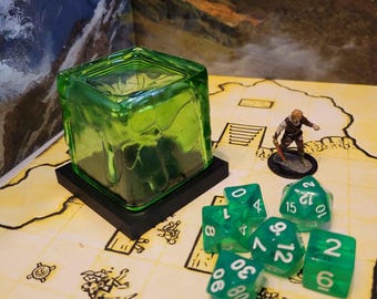 May include: A green glass cube with a textured surface sits on a black base.  The cube is surrounded by green dice with white numbers.  The numbers on the dice are 1, 2, 4, 6, 7, 8, 9, 12, 15, and 20.