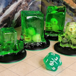 Op de afbeelding: Groene gelatine-kubussen met kleine witte schedels erin, op zwarte bases. Een groene 20-zijdige dobbelsteen bevindt zich op de voorgrond.