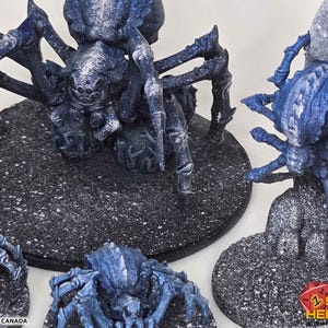 K&ouml;nnte beinhalten: Miniatur-Spinnenfiguren in Blau und Wei&szlig; mit detaillierten Texturen. Die Spinnen stehen auf strukturierten schwarzen Sockeln. Das Bild enth&auml;lt den Text "PRINTED IN CANADA" und das Shot Heroes-Logo.