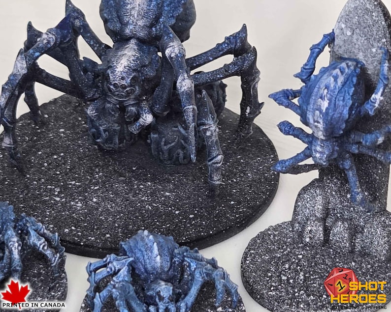 K&ouml;nnte beinhalten: Miniatur-Spinnenfiguren in Blau und Wei&szlig; mit detaillierten Texturen. Die Spinnen stehen auf strukturierten schwarzen Sockeln. Das Bild enth&auml;lt den Text "PRINTED IN CANADA" und das Shot Heroes-Logo.