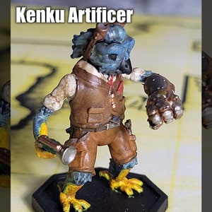 May include: A blue and brown painted miniature figurine of a Kenku Artificer, a bird-like humanoid with a mechanical arm, holding a tool in its other hand. The figurine is standing on a black and yellow base.