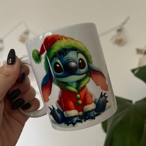 May include: White ceramic mug with a festive illustration of Stitch from Lilo & Stitch. The character is depicted in a Santa Claus outfit with a red coat and green hat. The mug has a white handle and is suitable for hot beverages.
