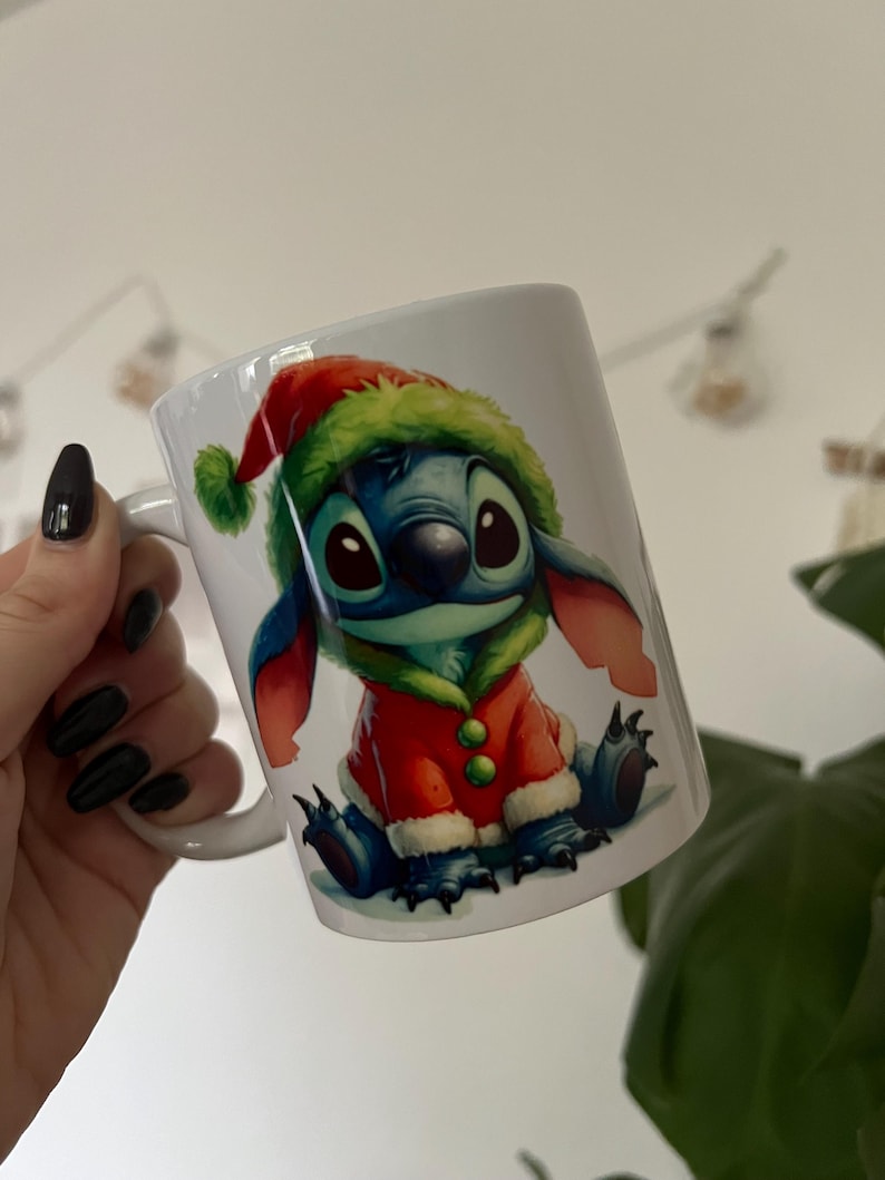 May include: White ceramic mug with a festive illustration of Stitch from Lilo & Stitch. The character is depicted in a Santa Claus outfit with a red coat and green hat. The mug has a white handle and is suitable for hot beverages.