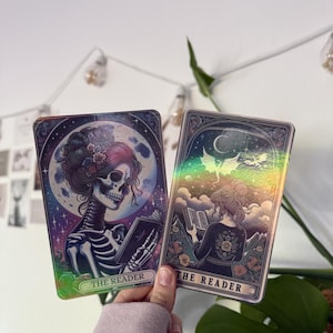 Könnte beinhalten: Zwei rechteckige Karten im Tarot-Stil mit der Aufschrift THE READER. Die linke Karte zeigt ein Skelett mit Blumenfrisur, das vor einem Vollmond ein Buch liest. Die rechte Karte zeigt eine Person von hinten, die unter einer Mondsichel ein Buch liest.