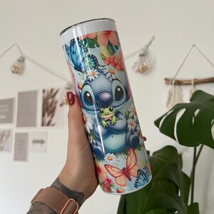 Puede incluir: Un vaso de acero inoxidable azul y blanco con un dise&ntilde;o floral que presenta a Stitch de Lilo & Stitch sosteniendo una rana verde. El vaso tiene una tapa blanca y una pajita.