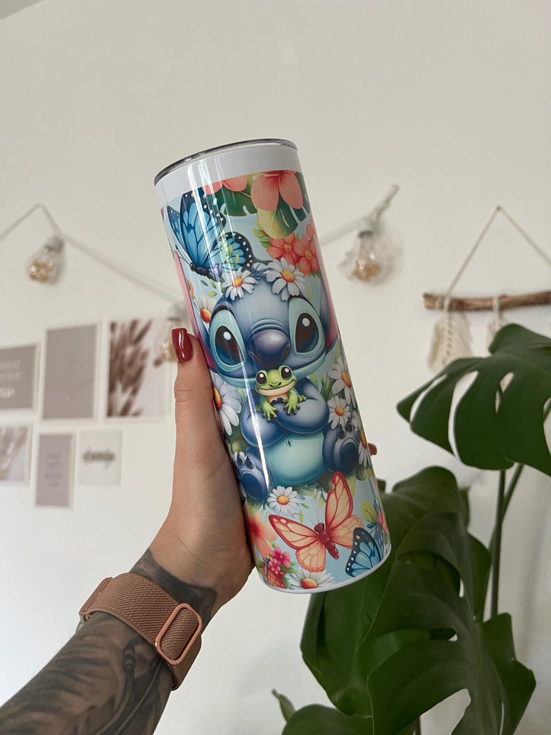 Puede incluir: Un vaso de acero inoxidable azul y blanco con un dise&ntilde;o floral que presenta a Stitch de Lilo & Stitch sosteniendo una rana verde. El vaso tiene una tapa blanca y una pajita.