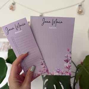 Puede incluir: Dos blocs de notas de papel de color morado claro con el nombre Jana Maria y la palabra Notizen impresos en la parte superior. Los diseños incluyen un patrón floral con flores rosas y blancas en la parte inferior y una cuadrícula.