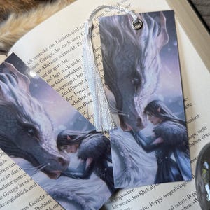 May include: Two rectangular bookmarks featuring a fantasy illustration of a person with long dark hair and pointed ears touching the snout of a large white and grey dragon. Each bookmark has a silver metal eyelet and a white silky tassel attached.