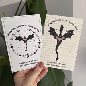 Può includere: Due biglietti d'auguri rettangolari con una silhouette di drago nero. Il testo recita: YOU WERE BORN TO RIDE DRAGONS AND CONQUER CHAOS. THE WORLD DOESN'T STAND A CHANCE. HAPPY BIRTHDAY, BONDED BADASS! Un biglietto ha un design a ghirlanda.