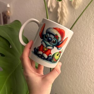 May include: White ceramic mug with a cartoon Stitch character dressed as Santa Claus. The design includes a red Santa hat, a red coat, and a green ornament. The mug has a curved handle and is held in a hand.
