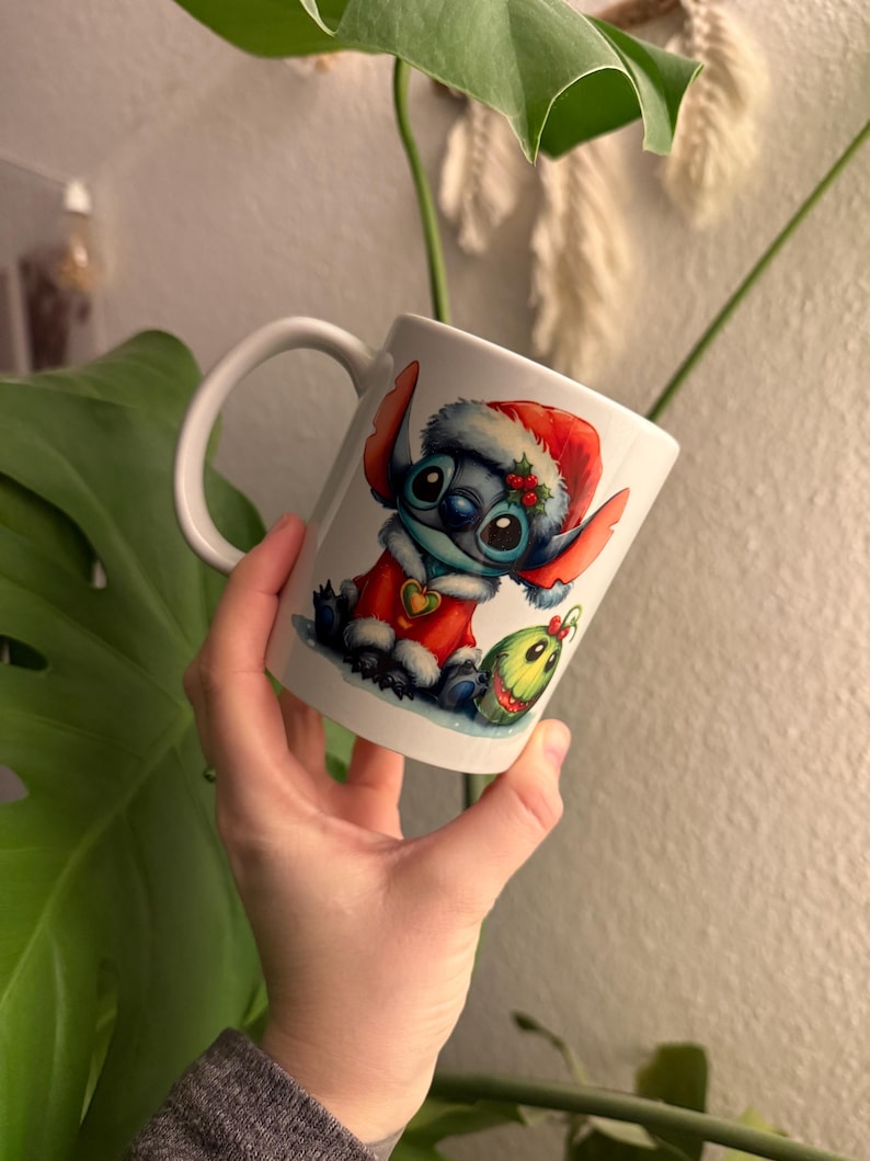 May include: White ceramic mug with a cartoon Stitch character dressed as Santa Claus. The design includes a red Santa hat, a red coat, and a green ornament. The mug has a curved handle and is held in a hand.