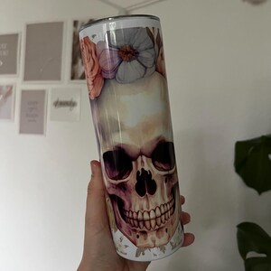 Puede incluir: Un vaso de acero inoxidable blanco con un dise&ntilde;o floral y un gr&aacute;fico de calavera. La calavera est&aacute; pintada en tonos de marr&oacute;n, rosa y morado.