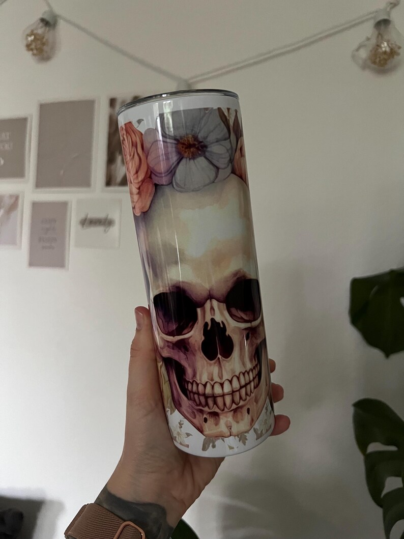 Puede incluir: Un vaso de acero inoxidable blanco con un dise&ntilde;o floral y un gr&aacute;fico de calavera. La calavera est&aacute; pintada en tonos de marr&oacute;n, rosa y morado.
