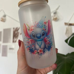 Könnte beinhalten: Ein mattes Glasgefäß mit einem Bambusdeckel, auf dem ein farbenfroher Comic-Charakter mit blauem Fell, großen Ohren und einer rosa Nase abgebildet ist. Der Charakter ist von Blumen und Wassertropfen umgeben.