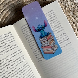 Könnte beinhalten: Ein blaues und weißes Lesezeichen mit einer Comicfigur mit blauem Fell und vier Fingern, die auf einem Stapel Bücher sitzt. Der Hintergrund ist hellblau mit weißen Sternen. Das Lesezeichen hat den Text "Der Mann, Scheibe Kuchen, ihren Teller, den noch mehr, aber es ihr in den mel fielen auf Alice, die hust"