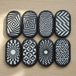 May include: Eight black and white patterned coasters with a black border. Each coaster features a unique geometric design, including stripes, squares, and starburst patterns. The coasters are arranged on a light brown wooden surface.