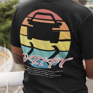 Op de afbeelding: Zwart T-shirt met een kleurrijke zonsondergang afbeelding van een auto die driften. De tekst "DRIFT" is in rood en wit onder de afbeelding gedrukt. De tekst onder de afbeelding luidt: "De neiging om grip te verliezen in de grenzen van de grip, waarbij een gecontroleerde overstuur in een overstuurconditie optreedt. De techniek werd in de jaren 1990 populair gemaakt door de Toyota Corolla en won aan populariteit bij veel bestuurders op de weg..."
