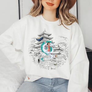Op de afbeelding: Wit sweatshirt met een grafische print van een meisje dat op een draak staat voor een Japanse tempel. De draak is wit met blauwe accenten. De tempel is in zwart-wit getekend.