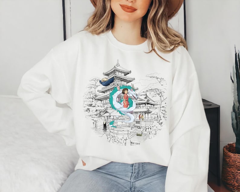 Op de afbeelding: Wit sweatshirt met een grafische print van een meisje dat op een draak staat voor een Japanse tempel. De draak is wit met blauwe accenten. De tempel is in zwart-wit getekend.