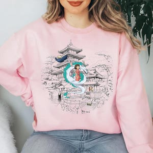 Op de afbeelding: Roze sweatshirt met een grafische print van een Japanse tempel, een draak en een meisje in een rode jurk. De grafiek is in zwart-wit met accenten in groen en blauw.