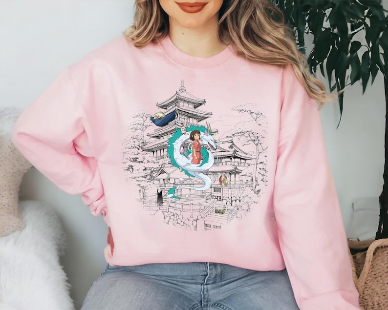 Op de afbeelding: Roze sweatshirt met een grafische print van een Japanse tempel, een draak en een meisje in een rode jurk. De grafiek is in zwart-wit met accenten in groen en blauw.