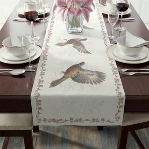 Puede incluir: Camino de mesa color crema con un diseño de faisanes en vuelo. El camino de mesa está decorado con un borde floral y está colocado sobre una mesa de madera oscura con cubiertos, copas de vino y un jarrón de flores rosas.