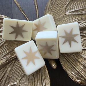 May include: Four rectangular off-white tiles, each featuring a centered metallic gold eight-pointed star design. The tiles are arranged in a cluster, resting upon a textured, metallic gold butterfly-shaped decorative object with detailed wing veins.