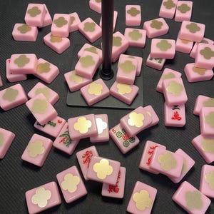 May include: Pink Mahjong tiles with gold clover designs and Chinese characters. The tiles are scattered across a dark surface. Some tiles have numbers and symbols. The tiles are a soft pink color.