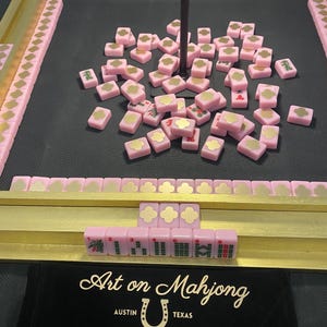 May include: A pink Mahjong set with gold accents. The set includes pink tiles with gold designs, gold racks, and a black carrying case with the text "Art on Mahjong Austin Texas". The tiles are scattered on a black surface.
