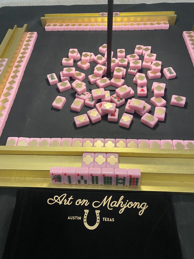May include: A pink Mahjong set with gold accents. The set includes pink tiles with gold designs, gold racks, and a black carrying case with the text "Art on Mahjong Austin Texas". The tiles are scattered on a black surface.