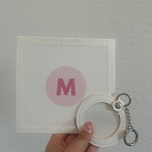 May include: A white canvas with a pink circle and the letter "M" in the center, next to a white leather keychain with a clear window and silver chain. The canvas has a woven texture.