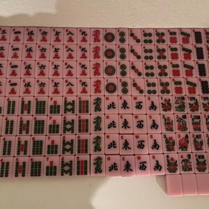 May include: A complete set of pink Mahjong tiles, arranged on a white surface. The tiles feature various symbols, numbers, and Chinese characters in red and black. The set includes tiles with circles, bamboo, and characters.