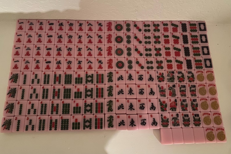 May include: A complete set of pink Mahjong tiles, arranged on a white surface. The tiles feature various symbols, numbers, and Chinese characters in red and black. The set includes tiles with circles, bamboo, and characters.