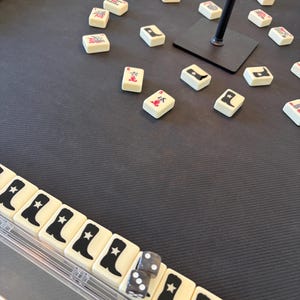 May include: A close-up of a Mahjong game in progress. The ivory-colored tiles feature black symbols and illustrations. Two dice are visible. The game is set on a dark gray surface.
