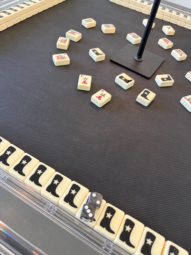May include: A Mahjong game in progress, with ivory tiles displaying black symbols and illustrations. Two dice are visible. The game is set on a dark grey surface.