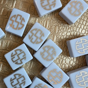 May include: A collection of white square tiles with a gold monogram design. The tiles are arranged on a textured gold surface. The monogram appears to be the letter "S" in a stylized circular pattern. The tiles are likely for a game or decorative use.