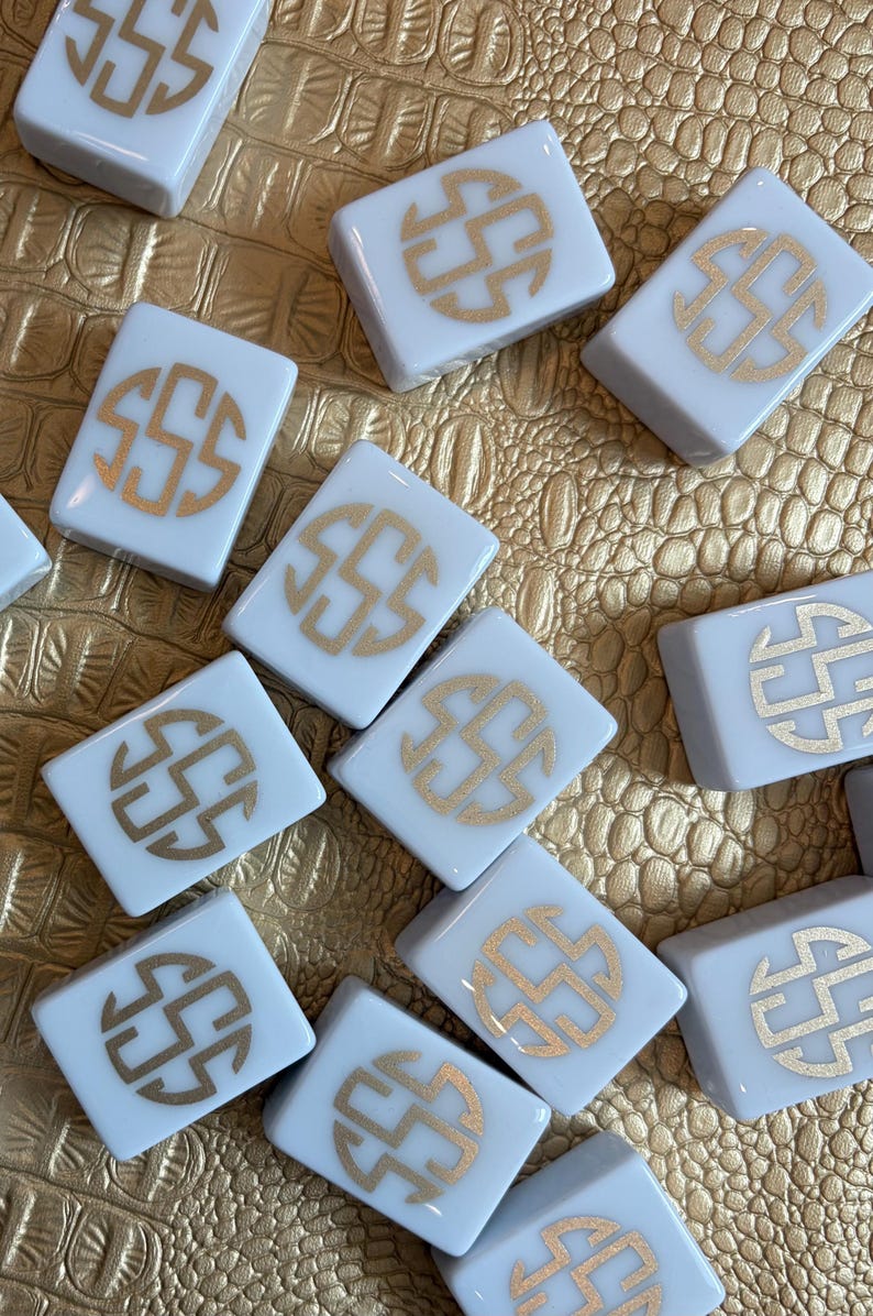 May include: A collection of white square tiles with a gold monogram design. The tiles are arranged on a textured gold surface. The monogram appears to be the letter "S" in a stylized circular pattern. The tiles are likely for a game or decorative use.