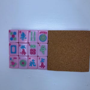 Pu&ograve; includere: Tessere di Mahjong rosa con vari simboli e numeri, tra cui la parola "Joker". Le tessere sono disposte accanto a una lavagna di sughero marrone. Le tessere presentano una variet&agrave; di colori, tra cui rosa, verde e blu.