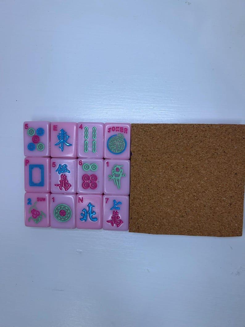 Pu&ograve; includere: Tessere di Mahjong rosa con vari simboli e numeri, tra cui la parola "Joker". Le tessere sono disposte accanto a una lavagna di sughero marrone. Le tessere presentano una variet&agrave; di colori, tra cui rosa, verde e blu.