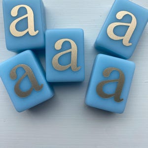 May include: Five light blue, square blocks with the lowercase letter "a" in gold. The blocks are arranged in a scattered pattern. The gold letter is centered on each block.