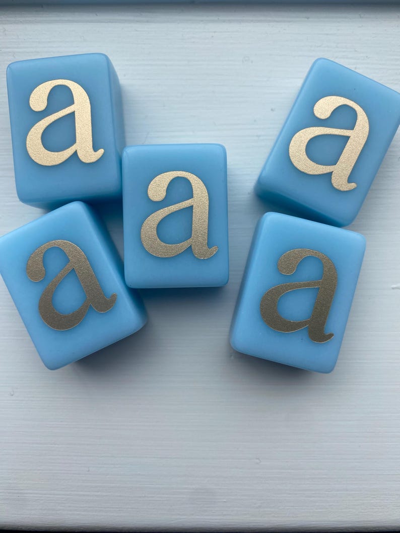 May include: Five light blue, square blocks with the lowercase letter "a" in gold. The blocks are arranged in a scattered pattern. The gold letter is centered on each block.