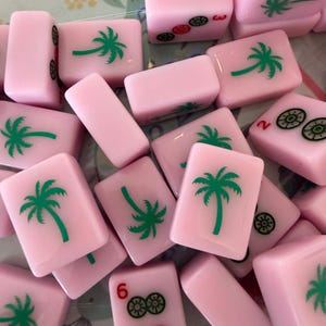May include: Pink Mahjong tiles with green palm tree and other symbols. The tiles are scattered, showing various numbers and symbols. The tiles are a soft pink color, and the palm trees are green.
